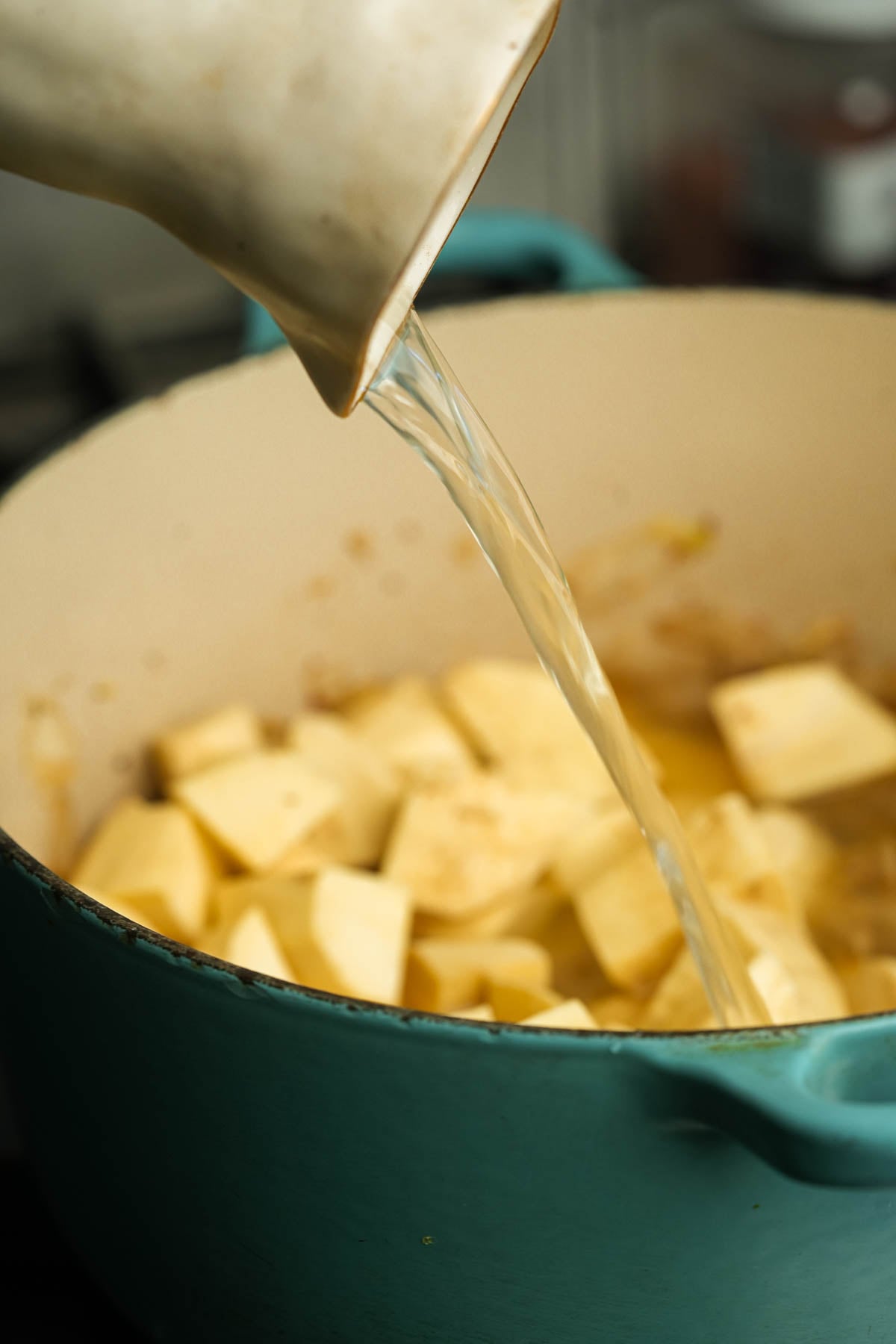 A white pitcher pours vegetable stock into a teal pot filled with chopped Japanese sweet potatoes.