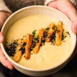 A person holds a bowl of creamy Japanese sweet potato soup topped with crispy fried strips, green onions, and a sprinkle of black and white sesame seeds.
