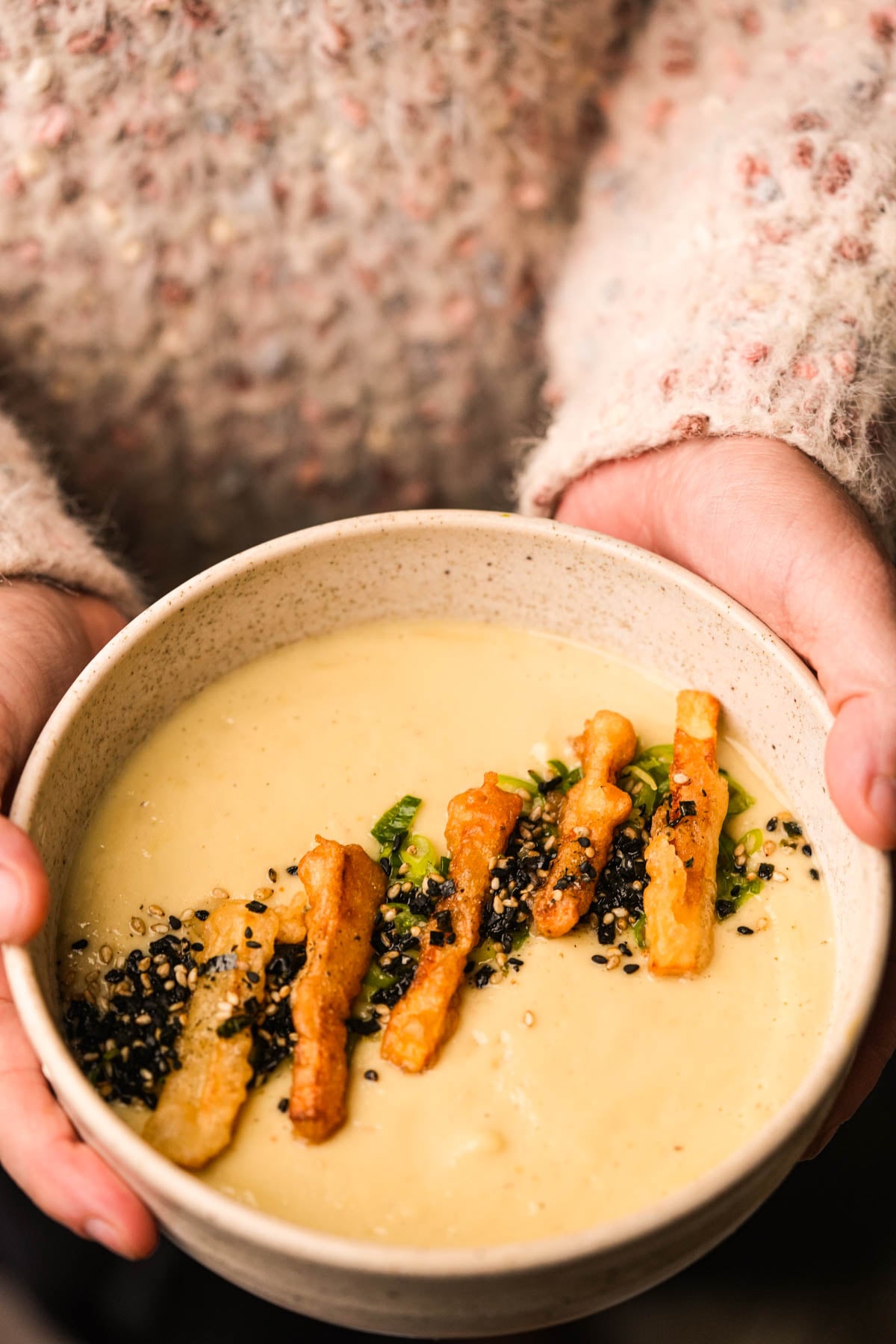 A person in a textured sweater holds a bowl of Japanese sweet potato soup topped with crispy tempura strips, sesame seeds, and chopped green onions.
