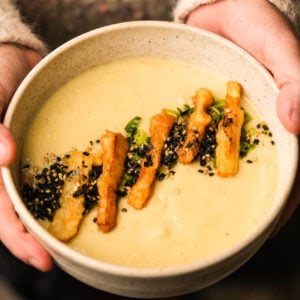 A person holds a bowl of creamy Japanese sweet potato soup topped with crispy fried strips, green onions, and a sprinkle of black and white sesame seeds.