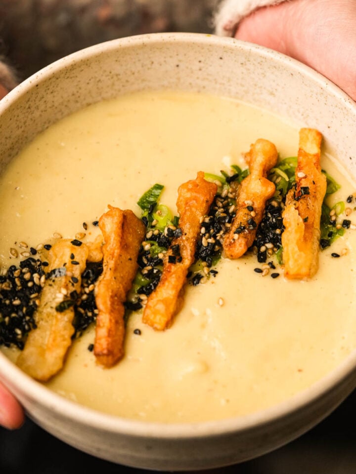 A person holds a bowl of creamy Japanese sweet potato soup topped with crispy fried strips, green onions, and a sprinkle of black and white sesame seeds.