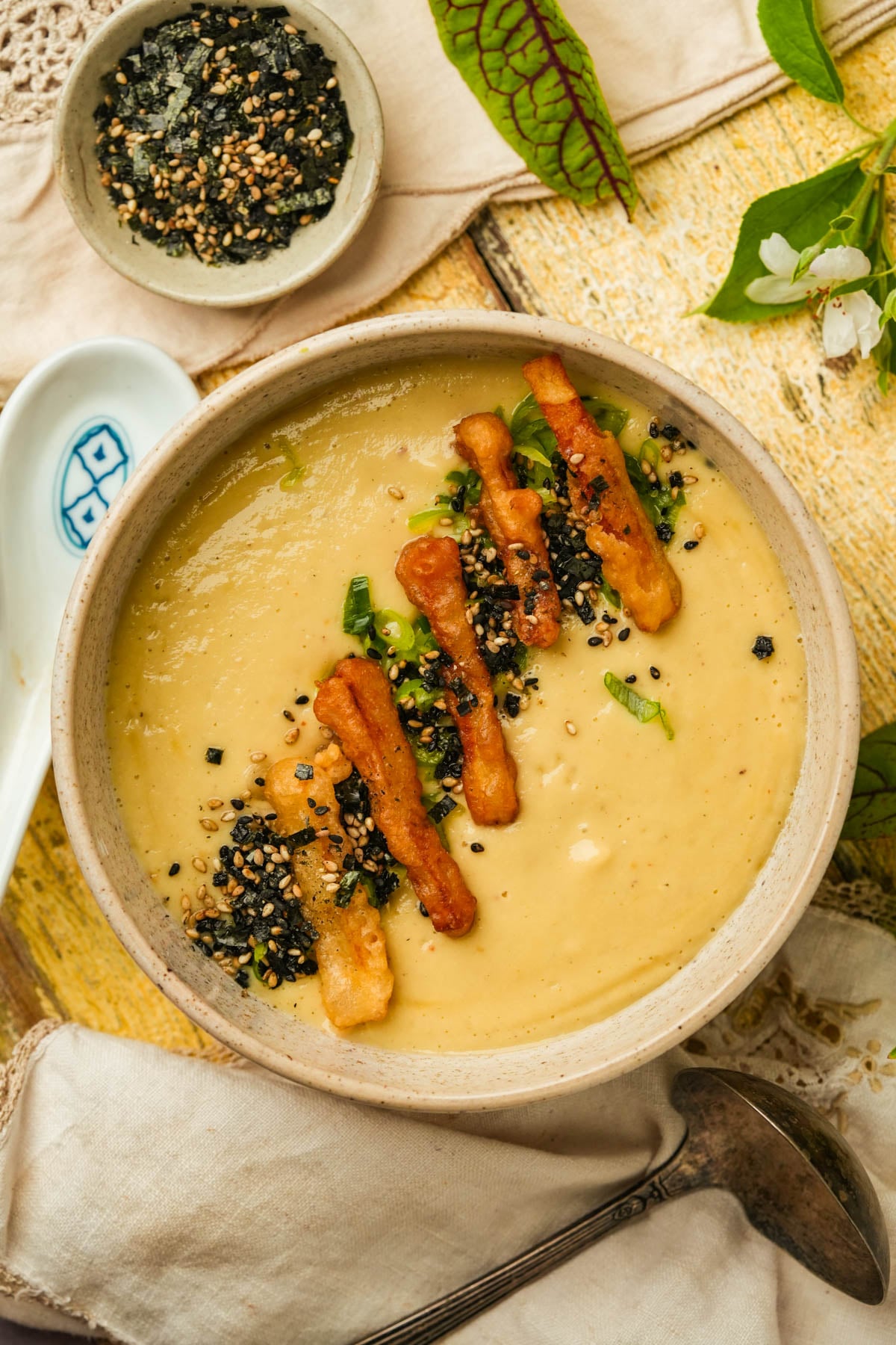 A bowl of creamy Japanese sweet potato soup topped with crispy strips, chopped green scallions, and sprinkled black sesame seeds, served on a beige cloth with a small dish of seeds nearby.