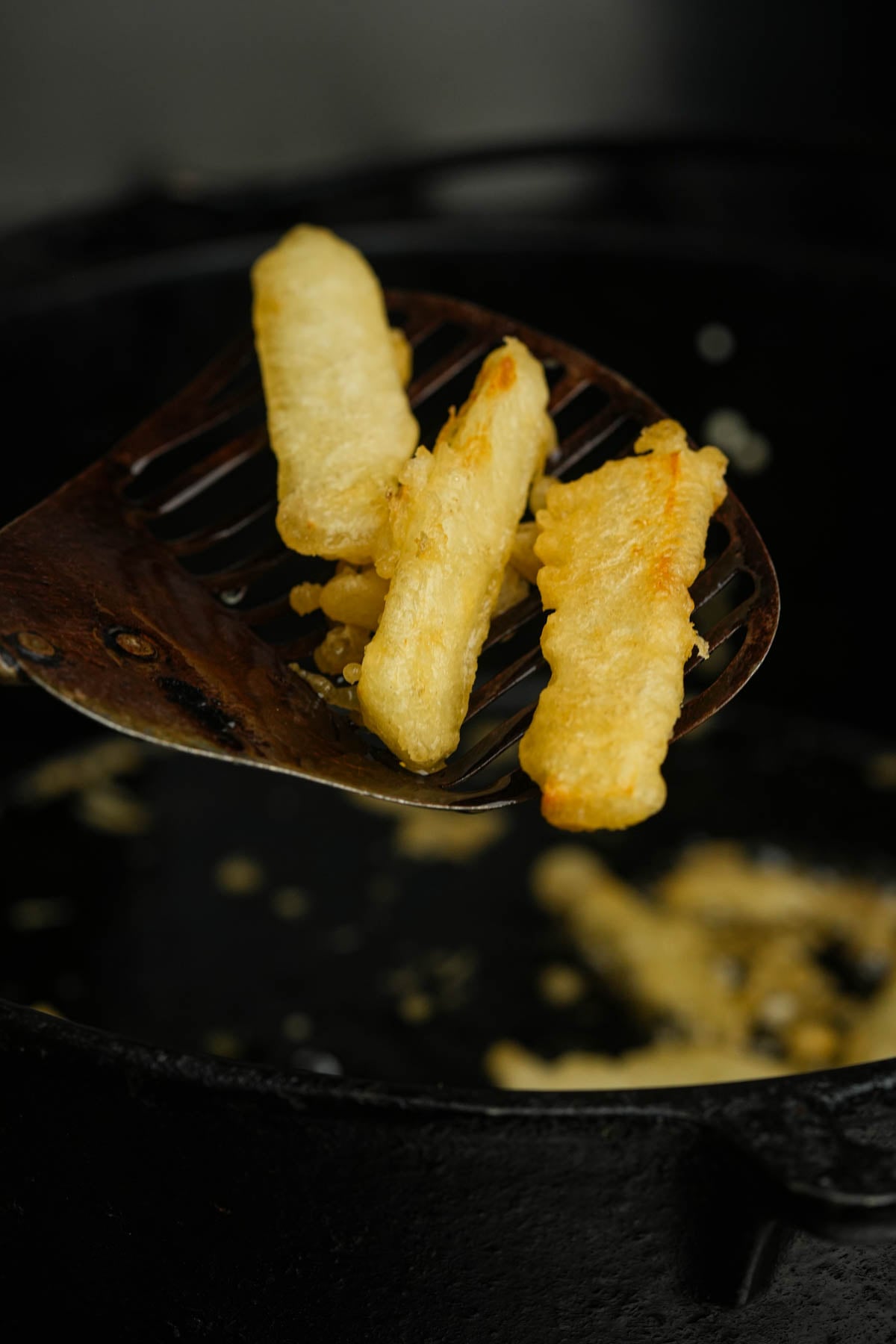 A slotted spatula holds three golden, crispy tempura sweet potatoes above a dark frying pan with oil and more strips cooking inside.
