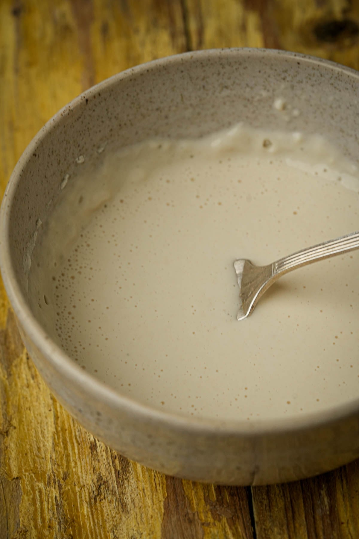 A beige ceramic bowl filled with a smooth, pale batter, with a silver fork resting inside. The bowl sits on a rustic, yellowish wooden surface.