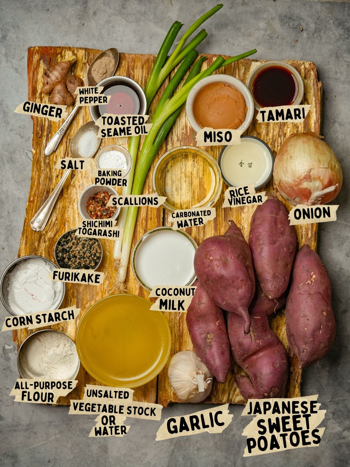 A wooden board topped with labeled ingredients for a Japanese sweet potato soup recipe: ginger, white pepper, toasted sesame oil, tamari, miso, rice vinegar, onion, scallions, salt, baking powder, shichimi togarashi, furikake, corn starch, flour, water, coconut milk, garlic, and Japanese sweet potatoes.