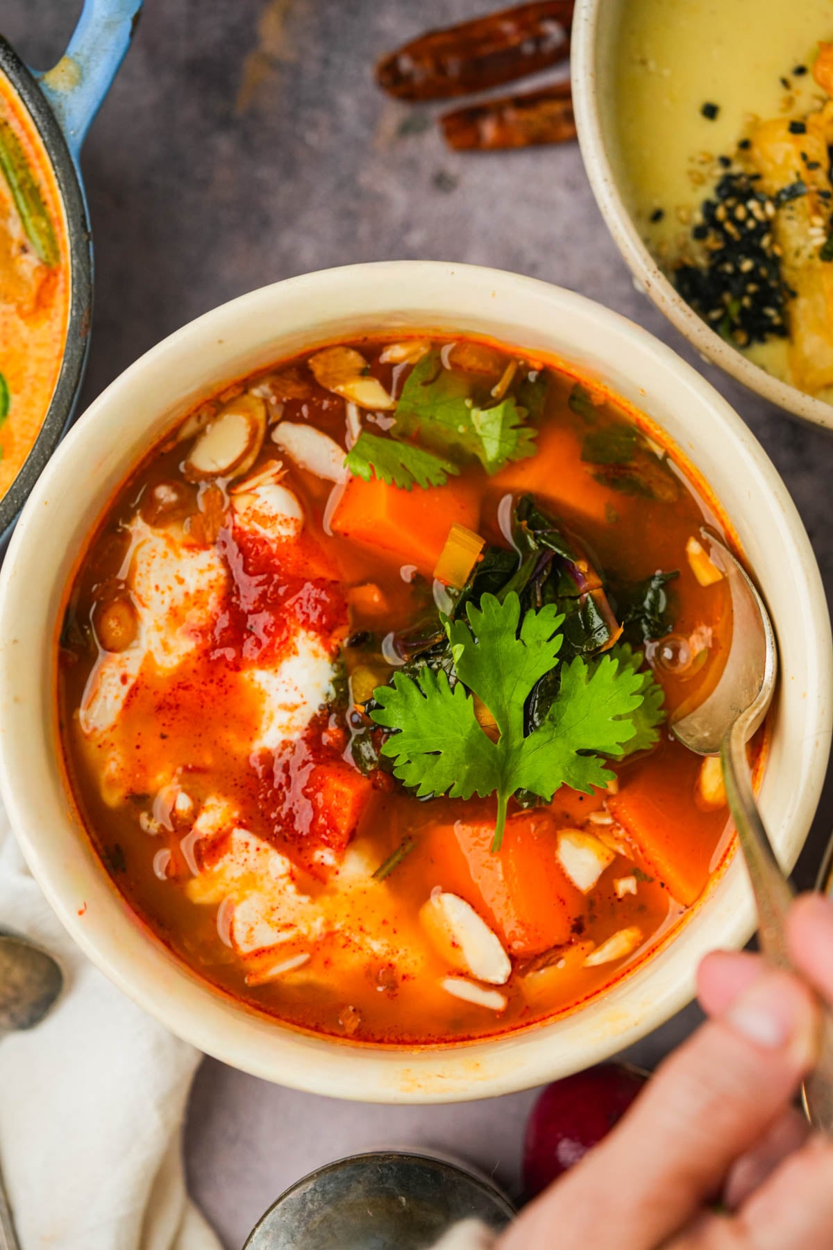 Bowl of Moroccan Sweet Potato Soup with tomato broth, cilantro garnish, and a dollop of cream. A hand holds a spoon in the bowl. Other dishes and utensils are partially visible around it.