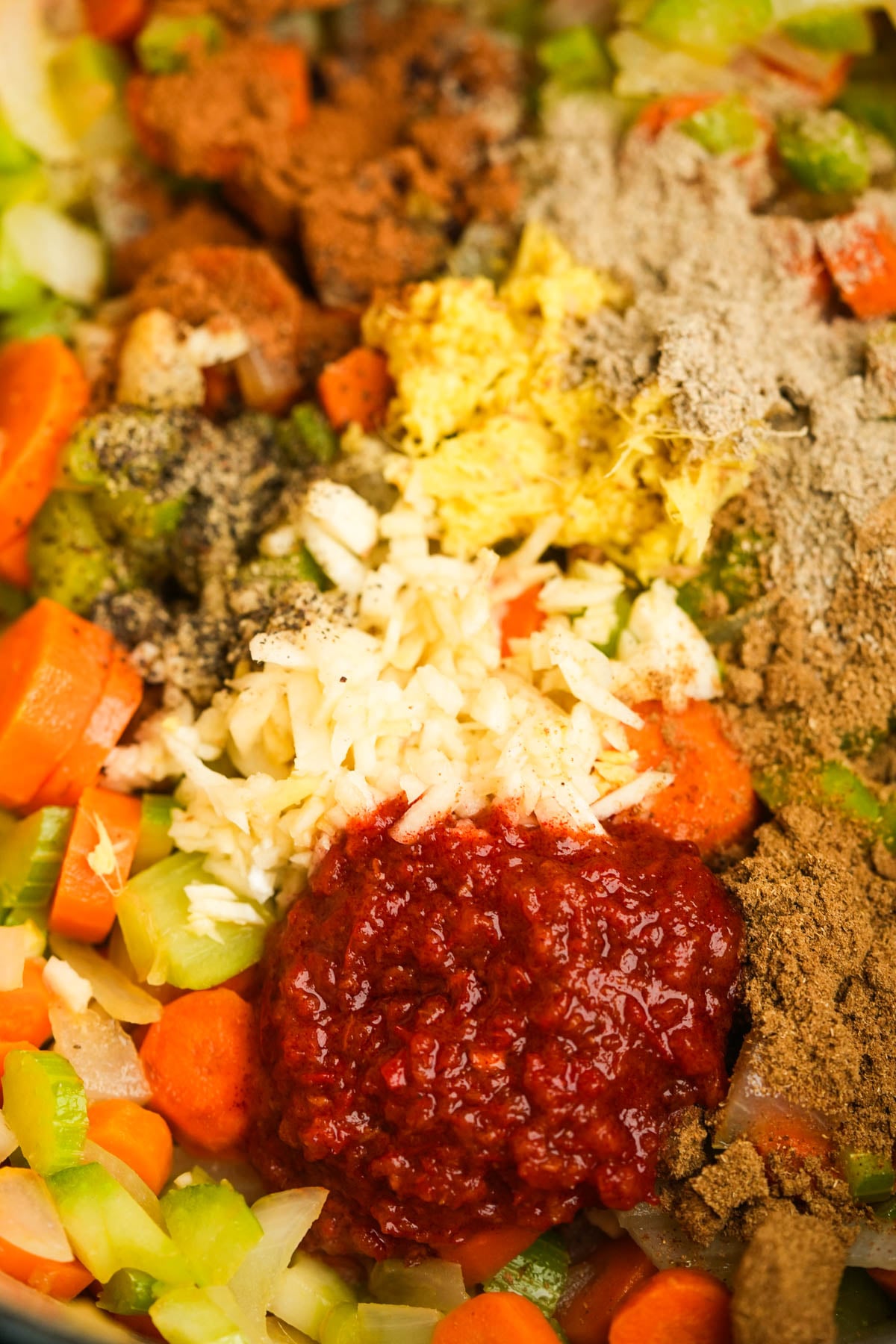 Close-up of chopped carrots, celery, onions, minced garlic, grated ginger, spices, and a dollop of harissa paste mixed together, ready to be cooked.