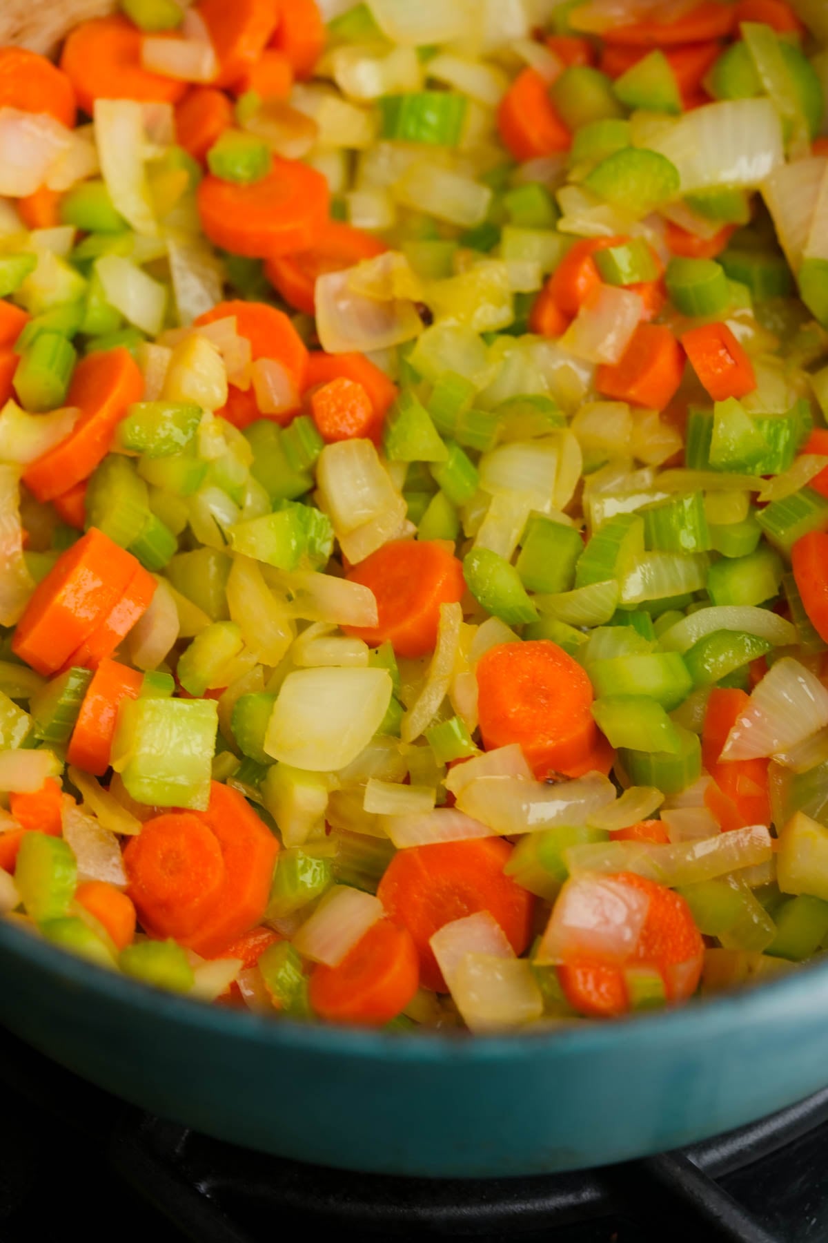 Chopped carrots, celery, and onions are sautéing in a pan. The vegetables are cut into small pieces and appear to be cooking, with vibrant orange, green, and white colors visible.