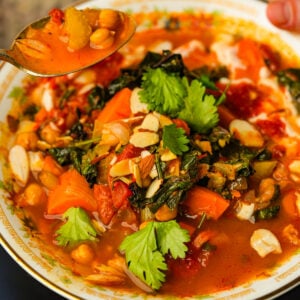 A colorful Moroccan sweet potato soup with chickpeas, carrots, greens, tomato broth, sliced almonds, and fresh cilantro served in a bowl, with a spoonful lifted above.