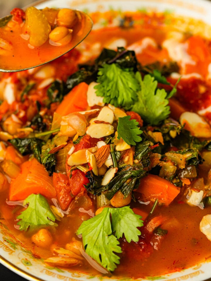 A colorful Moroccan sweet potato soup with chickpeas, carrots, greens, tomato broth, sliced almonds, and fresh cilantro served in a bowl, with a spoonful lifted above.