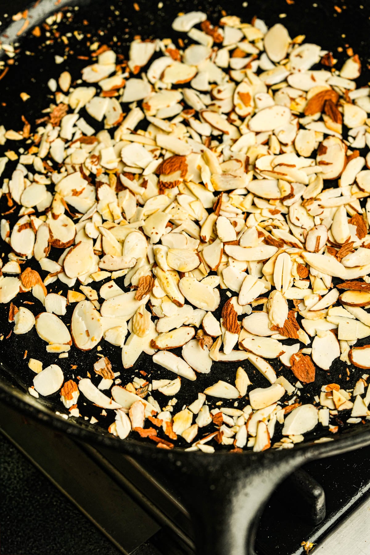 Sliced almonds being toasted in a black cast iron skillet on a stovetop.