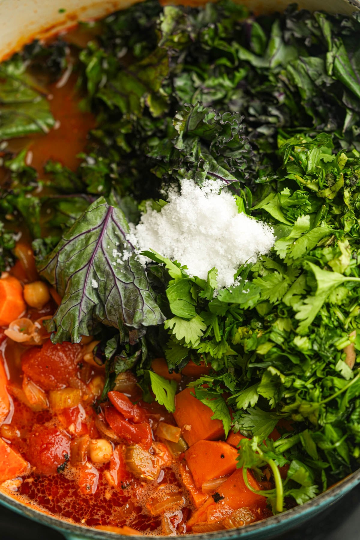 A a pot of Moroccan sweet potato soup with leafy greens, chopped cilantro, a pinch of salt, chickpeas, tomatoes, and sliced carrots in a rich broth.