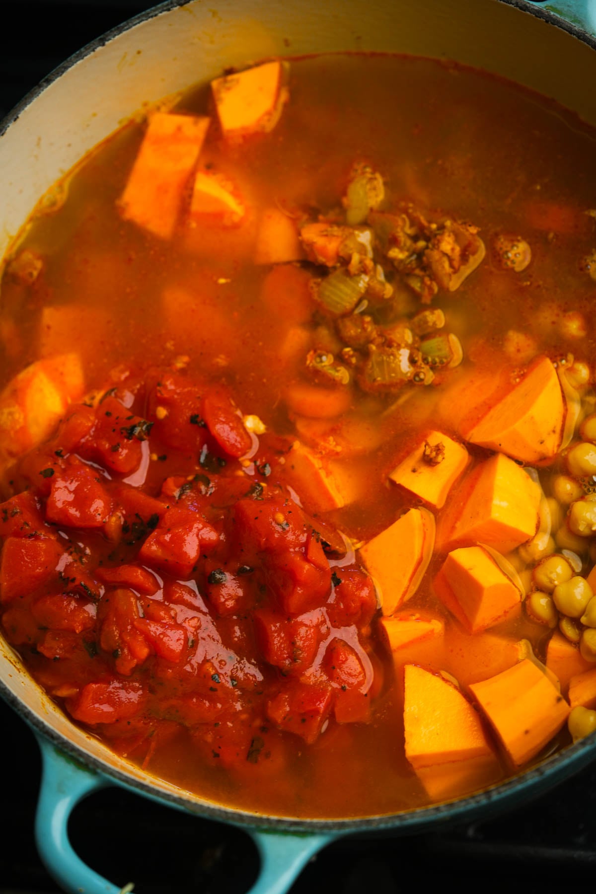A pot of Moroccan sweet potato soup with diced sweet potatoes, chickpeas, celery, and chopped tomatoes in a seasoned broth, cooking on a stovetop.