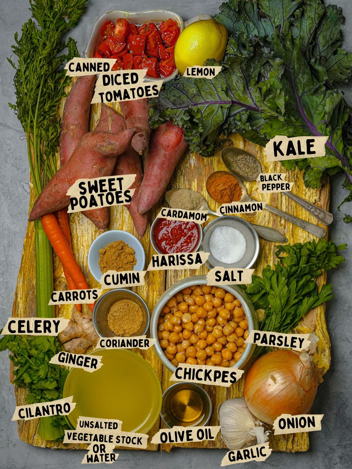A wooden board with labeled ingredients for a harissa sweet potato soup: sweet potatoes, canned diced tomatoes, lemon, kale, carrots, celery, cilantro, ginger, vegetable stock, olive oil, chickpeas, parsley, onion, garlic, cumin, coriander, harissa, cinnamon, salt, and black pepper.