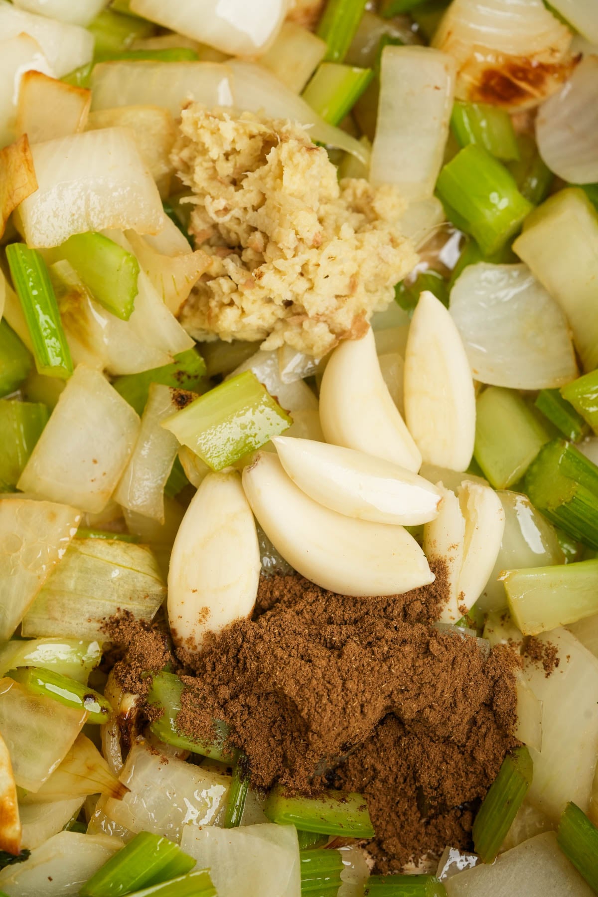 Chopped onions and celery are sautéed in a pan with whole garlic cloves, minced ginger, and ground spices, ready to be mixed together.