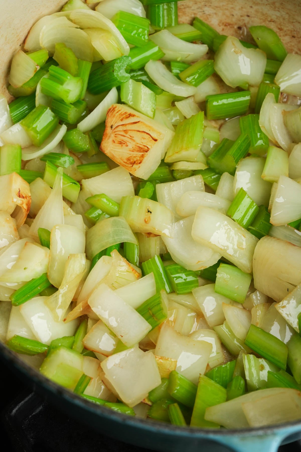 Chopped onions and celery sautéing in a pan, with some pieces lightly browned. The vegetables are glistening, indicating they are being cooked in oil.