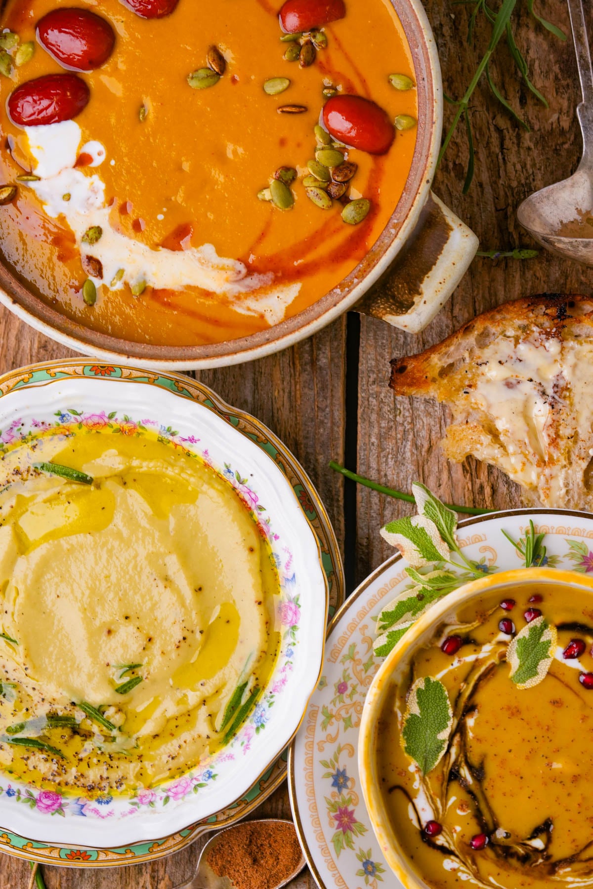 Three colorful bowls of creamy soup, garnished with herbs, seeds, and sauces, sit on a rustic wooden table beside a slice of bread.