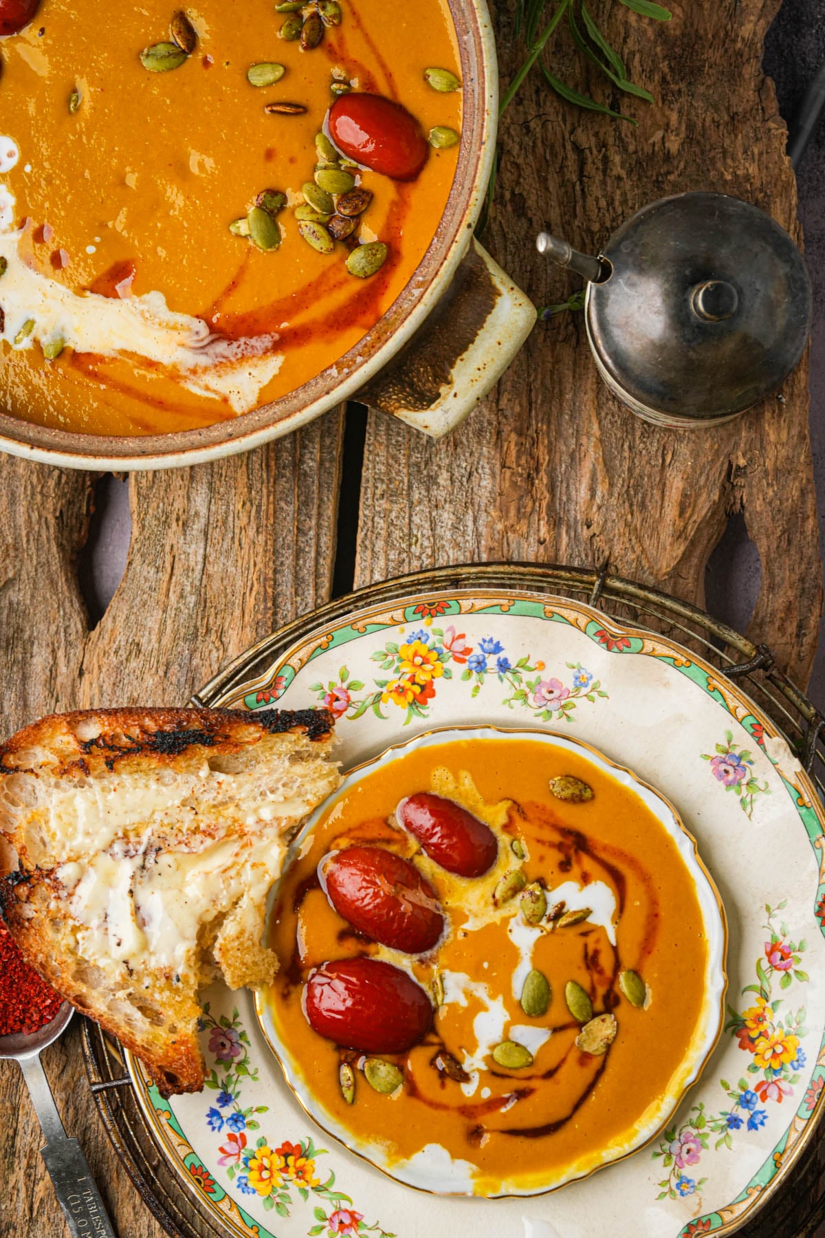 Bowl of creamy kabocha squash soup with jujube dates, pumpkin seeds, and coconut milk served with toasted bread on a rustic floral plate atop a wooden table.