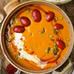 A bowl of creamy kabocha squash soup topped with red jujube dates, pumpkin seeds, and a swirl of coconut milk set on a rustic wooden table.