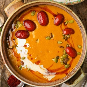 A bowl of creamy kabocha squash soup topped with red jujube dates, pumpkin seeds, and a swirl of coconut milk set on a rustic wooden table.