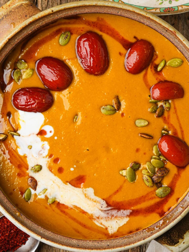 A bowl of creamy kabocha squash soup topped with red jujube dates, pumpkin seeds, and a swirl of coconut milk set on a rustic wooden table.