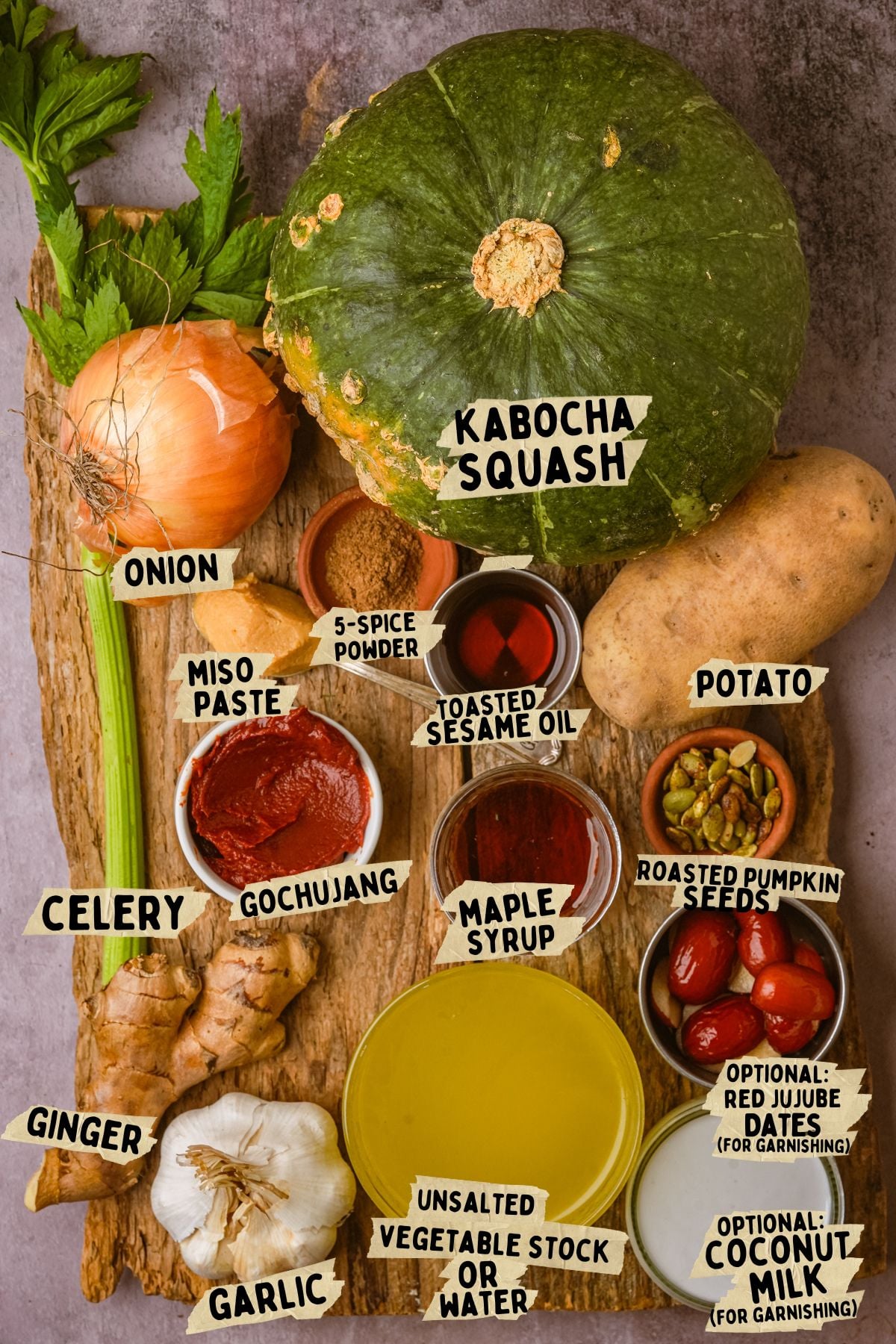 A wooden board with ingredients for kabocha squash soup recipe: kabocha squash, onion, potato, celery, ginger, garlic, miso paste, gochujang, 5-spice powder, sesame oil, maple syrup, broth, roasted pumpkin seeds, jujube dates, and coconut milk.