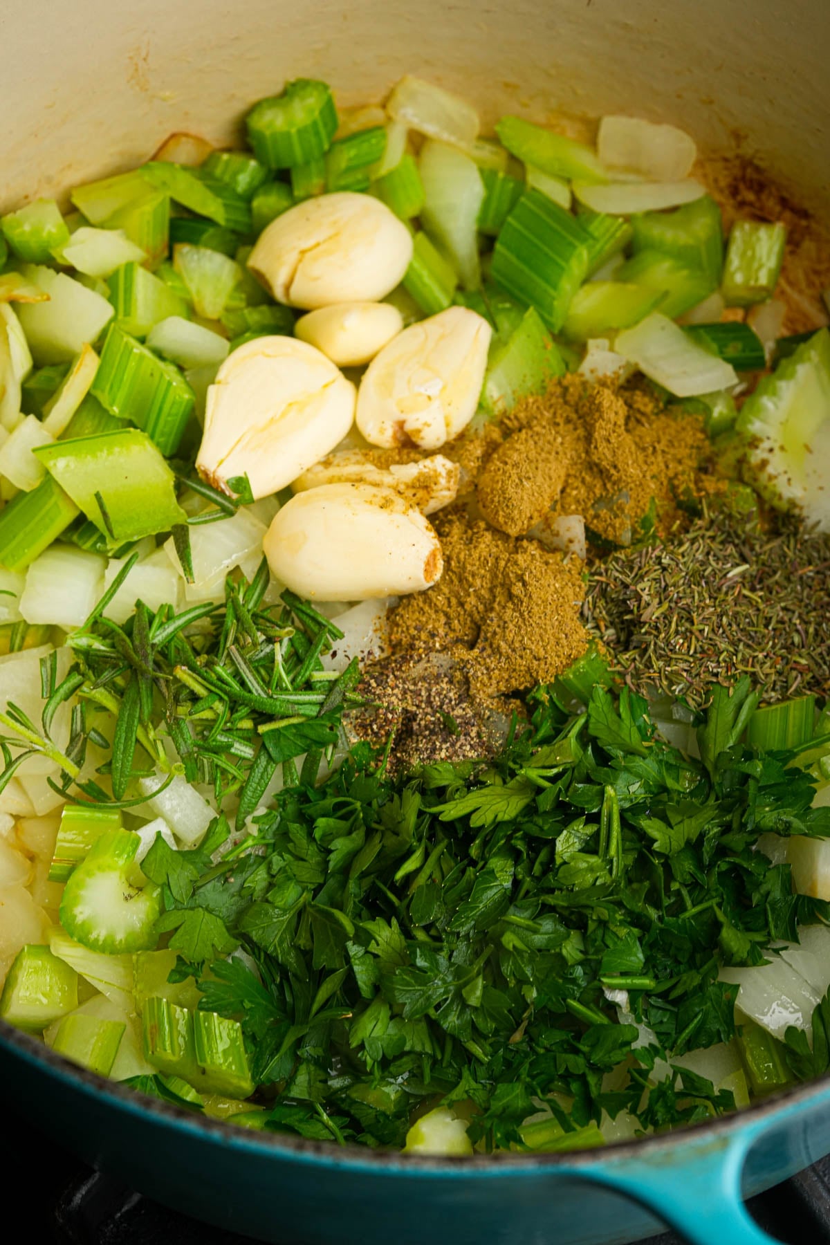 Chopped celery, onions, parsley, whole garlic cloves, rosemary, thyme, ground spices, and black pepper in a blue pot.