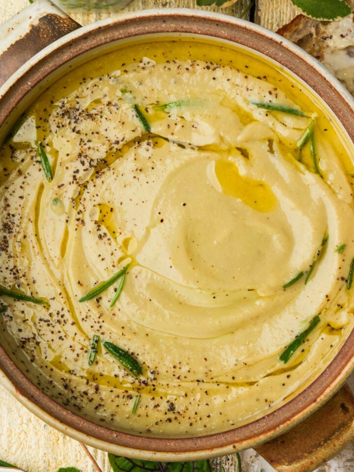A bowl of vegan truffle cauliflower soup is garnished with olive oil, fresh rosemary, and black pepper.