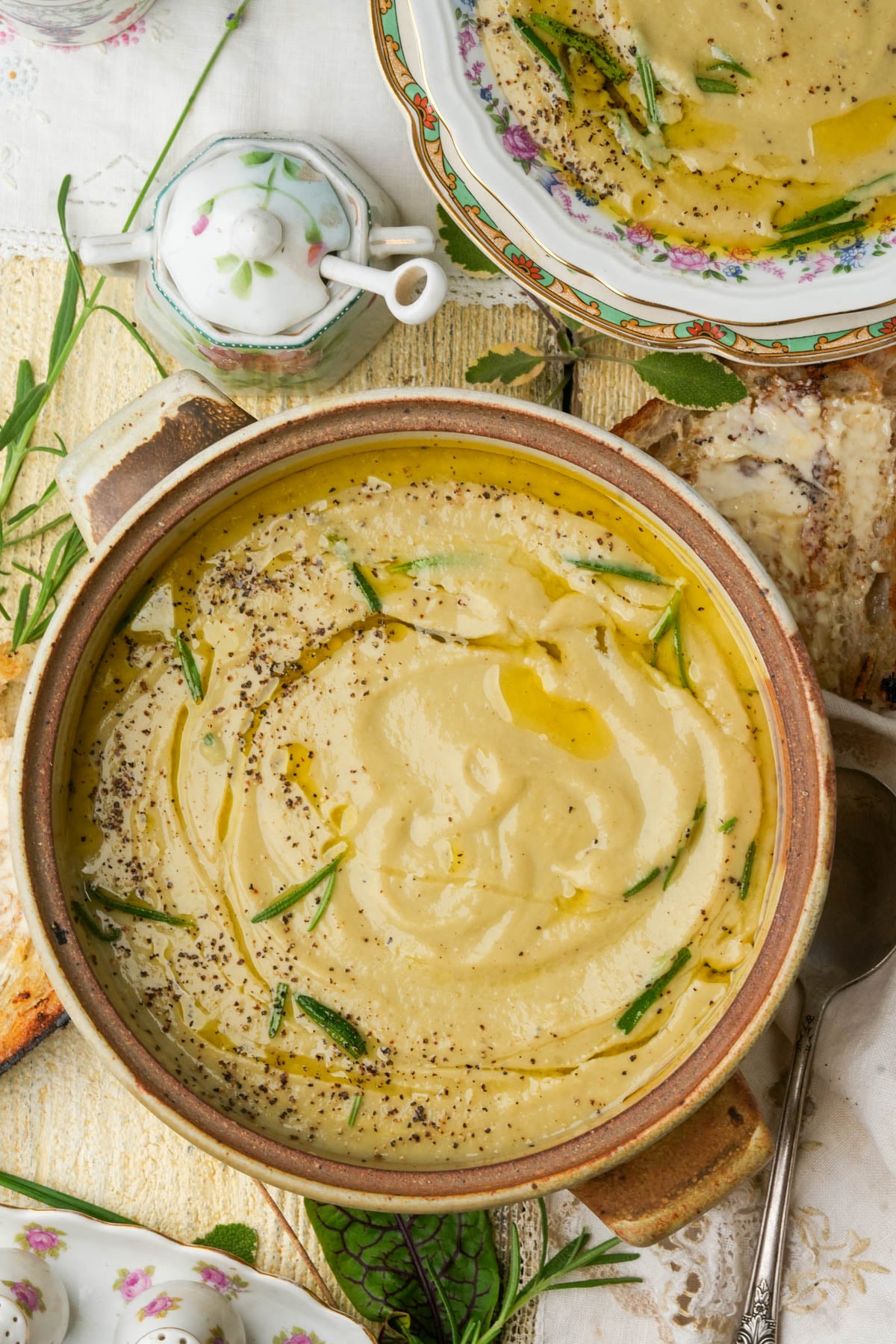 Vegan truffle cauliflower soup garnished with herbs, black pepper, and a drizzle of olive oil in a brown ceramic bowl. A spoon, toast, and a floral china plate with more soup are beside it.