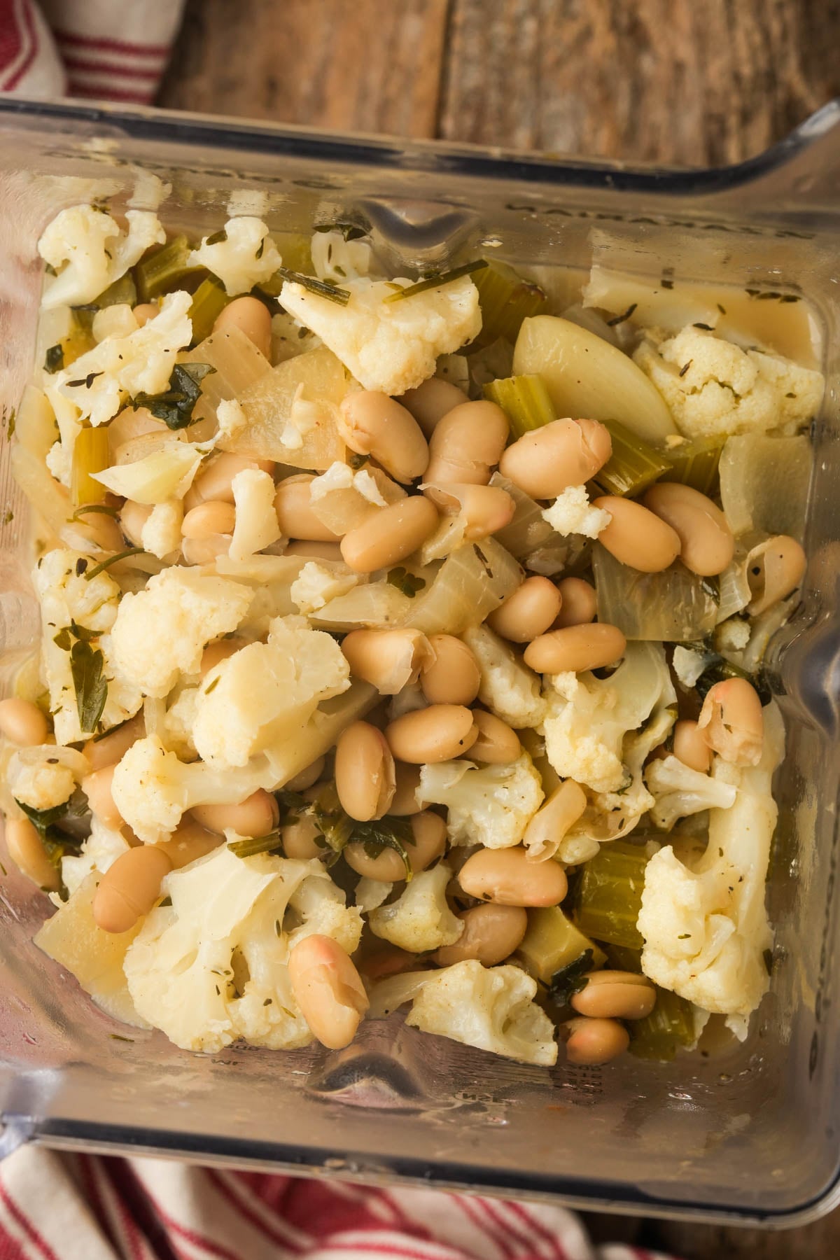 Chopped cauliflower, white beans, onions, and herbs in a blender, ready for blending. Some ingredients are roughly chopped. The container is sitting on a wooden surface with a striped cloth nearby.