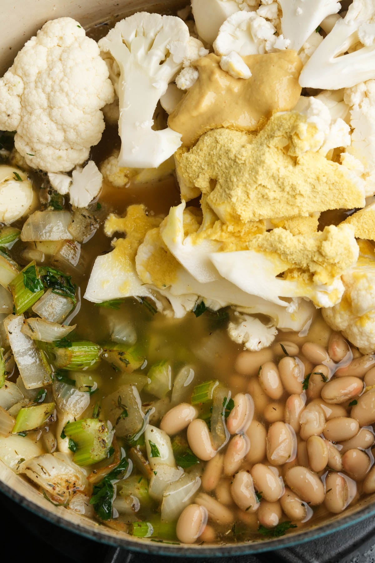 A close-up of a pot containing chopped onions, celery, cooked white beans, cauliflower florets, vegetable broth, and a large scoop of nutritional yeast and mustard.