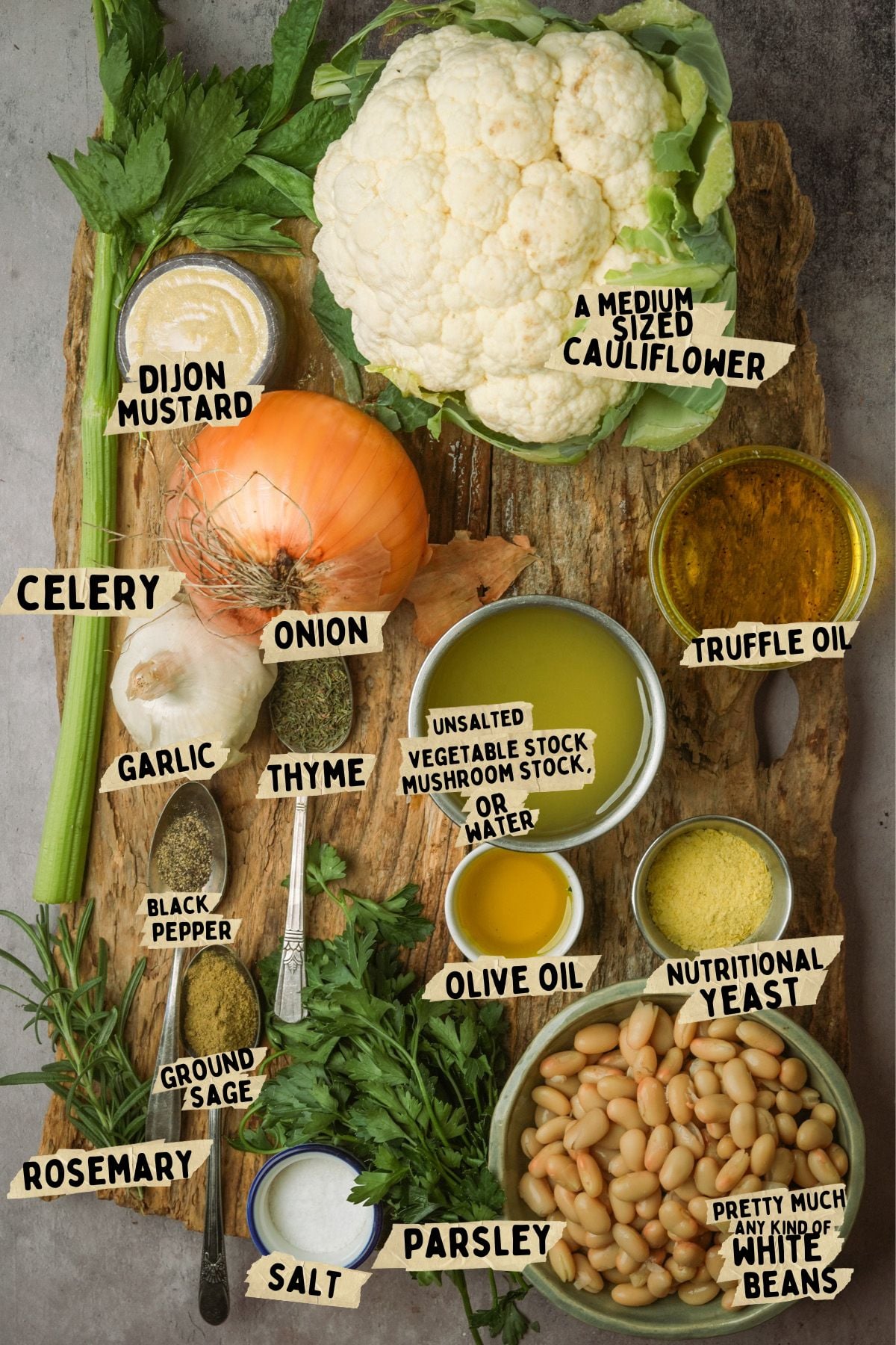 A variety of fresh vegetables, herbs, and ingredients for a truffle cauliflower soup recipe are arranged on a table, including cauliflower, celery, onion, olive oil, garlic, rosemary, white beans, and small bowls of seasoning and spices.