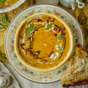 A bowl of creamy acorn squash soup garnished with sage leaves, pomegranate seeds, cream, and a swirl of coconut milk, served on a floral plate with a slice of buttered bread on the side.