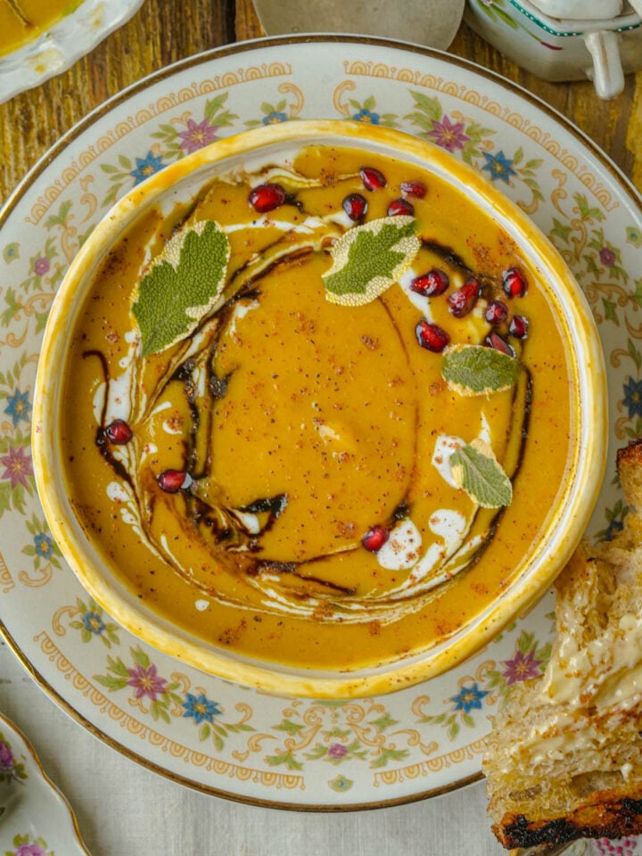 A bowl of creamy acorn squash soup garnished with sage leaves, pomegranate seeds, cream, and a swirl of coconut milk, served on a floral plate with a slice of buttered bread on the side.