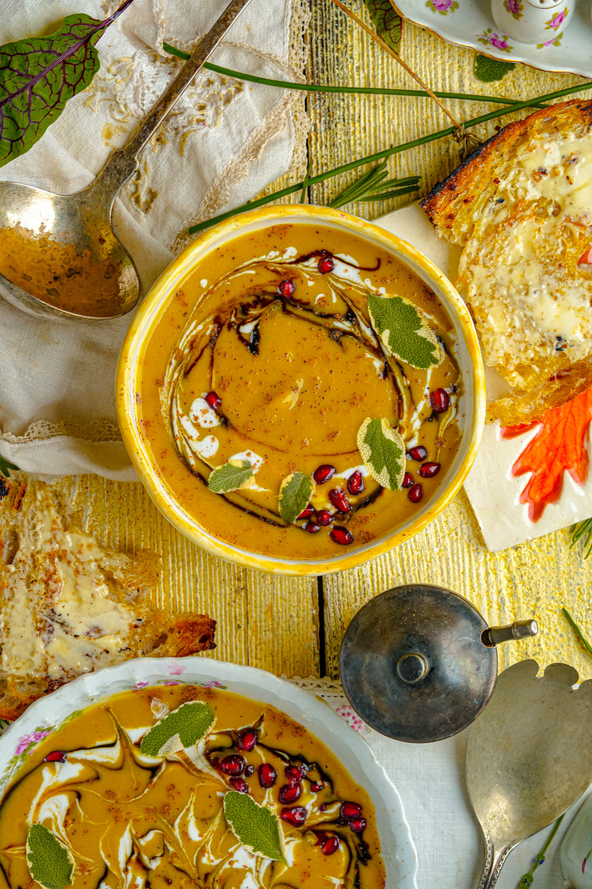 A bowl of vegan acorn squash soup garnished with herbs and pomegranate seeds, surrounded by buttered toast, vintage silverware, and a floral napkin on a rustic yellow wooden table.
