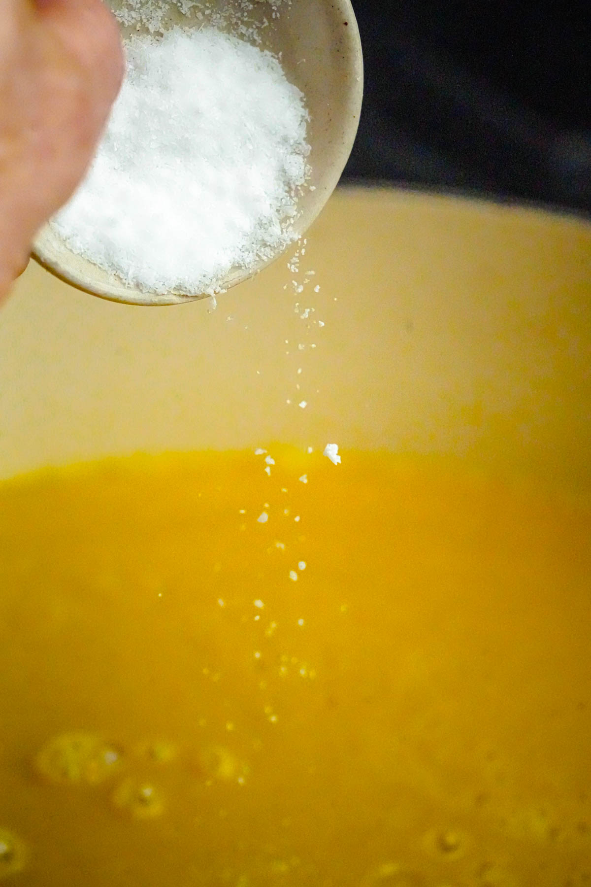 A hand sprinkles salt from a small bowl into a pot containing acorn squash soup.