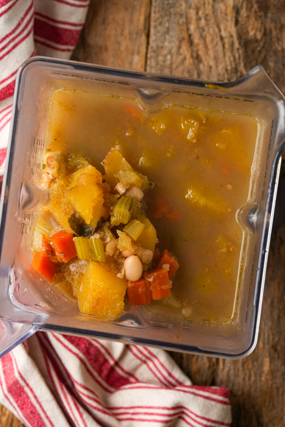 A blender filled with unblended acorn squash soup, including carrots, celery, beans, and squash, sitting on a rustic wooden surface next to a red-striped towel.
