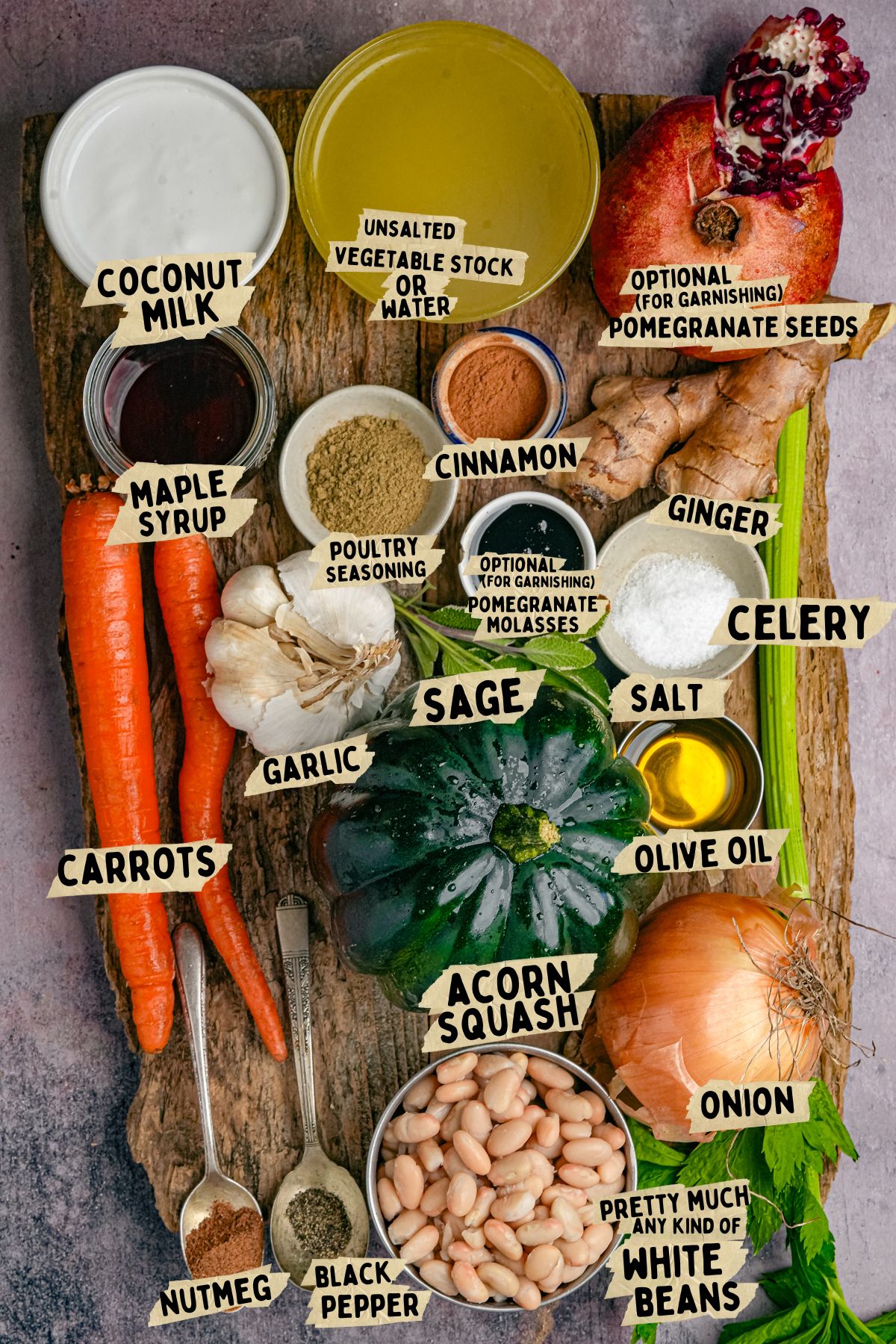 Labeled ingredients for a acorn squash soup recipe on a wooden board: coconut milk, maple syrup, carrots, garlic, nutmeg, black pepper, unsalted vegetable stock, cinnamon, sage, acorn squash, white beans, onion, celery, olive oil, ginger, pomegranate seeds.