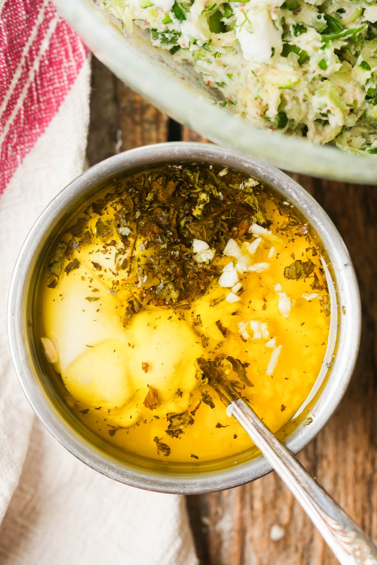 A metal bowl filled with oil, herbs, vegan yogurt, lemon juice, and minced garlic, with a spoon inside.
