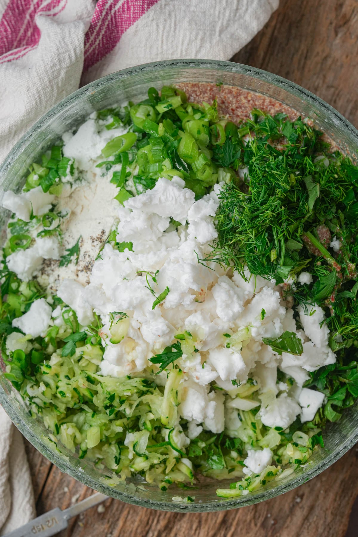 A glass bowl containing chopped green onions, fresh herbs, grated zucchini, crumbled feta cheese, and flour, all ready to be mixed to become mukver batter. A pink-striped towel is partially visible.