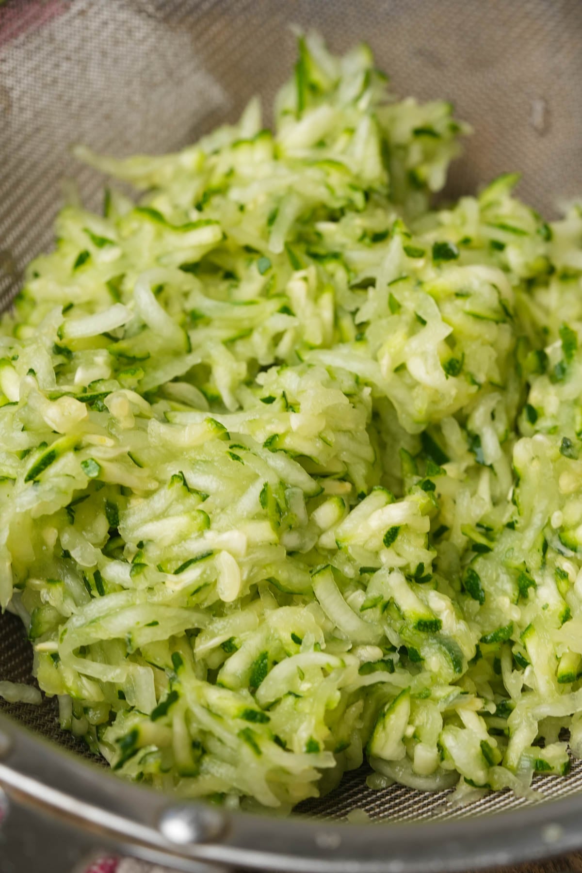 Shredded zucchini rests in a metal mesh strainer, showing green and white strands with visible moisture, ready for use in cooking or baking.
