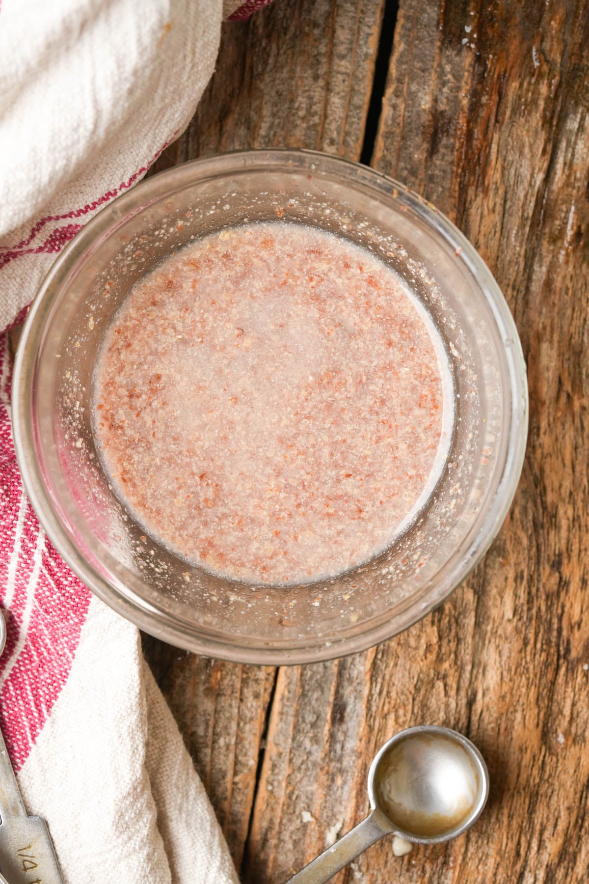 A glass bowl with a mixture of ground flaxseed and water sits on a rustic wooden surface next to a metal measuring spoon and a white towel with red stripes.