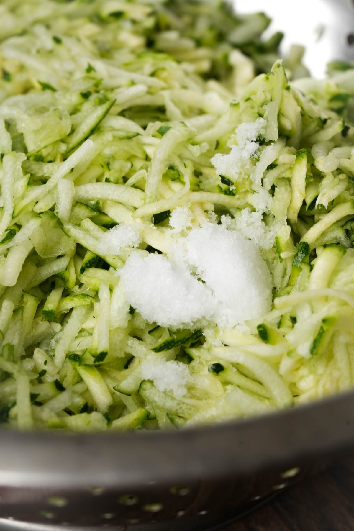 Close-up of shredded zucchini with a small pile of coarse salt sprinkled on top, sitting in a metal colander.