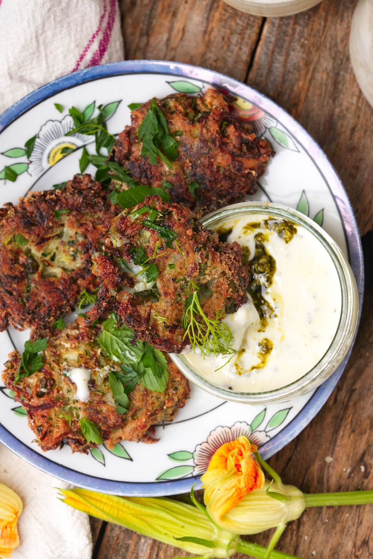 Three zucchini fritters garnished with herbs on a floral plate, served with a small jar of creamy dipping sauce swirled with green pesto. A squash blossom and napkin are nearby on a rustic wooden table.