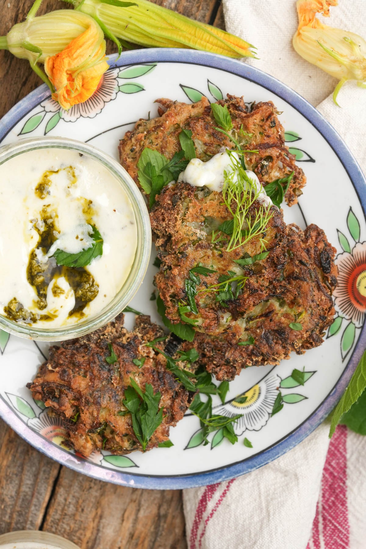 Three crispy mukver fritters garnished with herbs sit on a floral plate beside a bowl of creamy dipping sauce with green pesto. Edible zucchini flowers and a cloth napkin decorate the rustic wooden table.