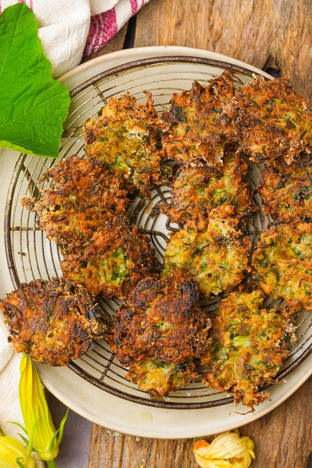 A plate of golden-brown zucchini fritters sits on a round wire rack, garnished with green leaves and zucchini blossoms, atop a rustic wooden surface.