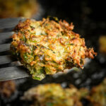 A close-up of a golden-brown mukver fritter with crispy edges, held by a metal spatula above a dark, blurred background.