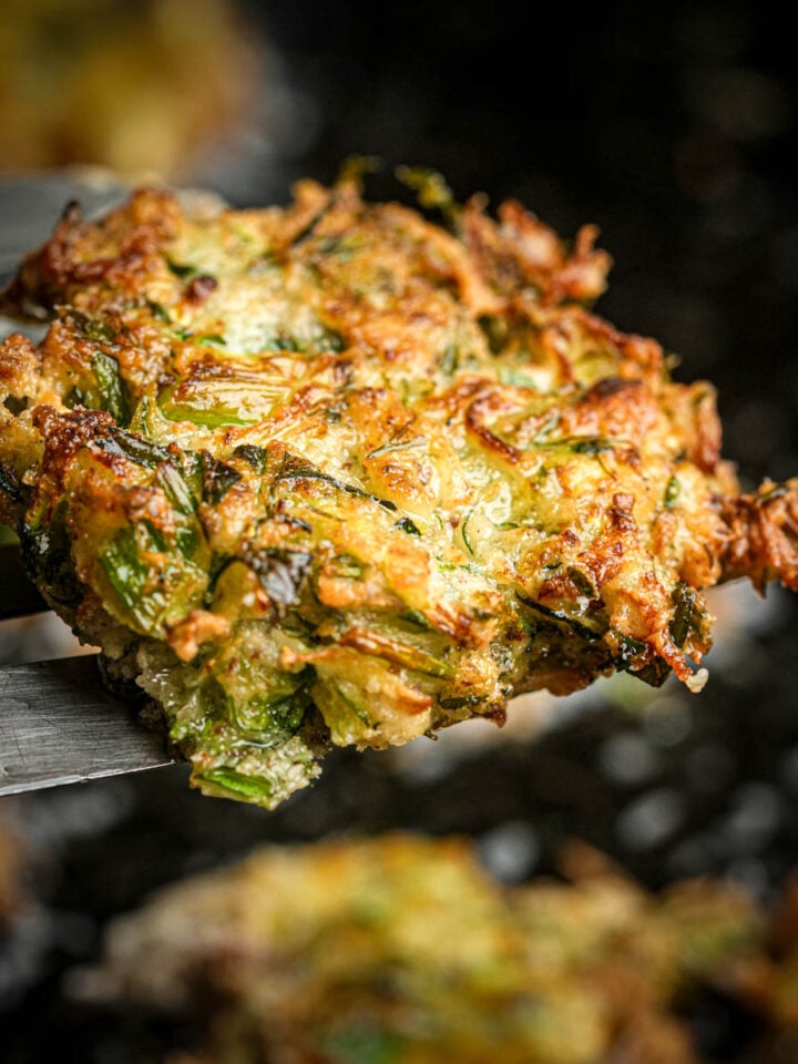 A close-up of a golden-brown mukver fritter with crispy edges, held by a metal spatula above a dark, blurred background.