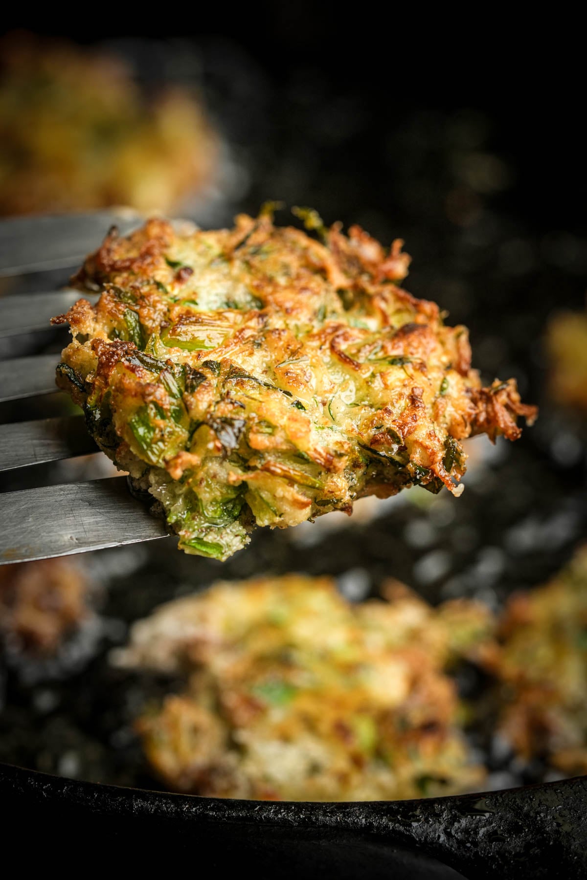 A metal spatula lifts a crispy, golden-brown mukver fritter with visible herbs from a dark pan, with more fritters blurred in the background.