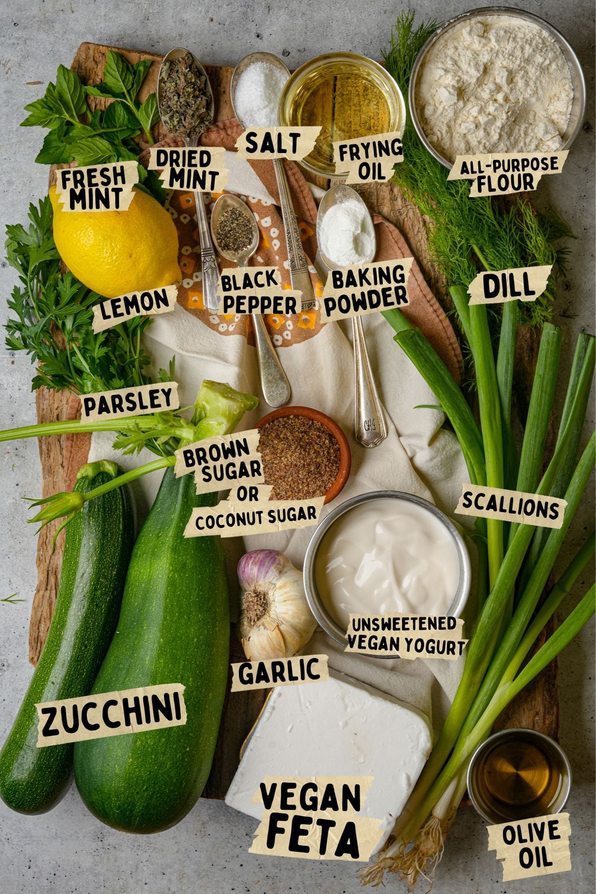 Ingredients for a vegan mukver recipe arranged on a wooden board, including zucchini, parsley, dill, scallions, garlic, lemon, vegan feta, yogurt, oil, flour, baking powder, mint, salt, pepper, and sugar.